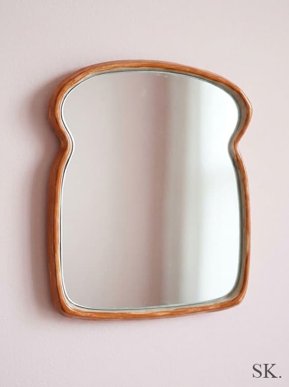 A whimsical mirror shaped like a slice of bread with a brown crust-like frame, hanging on a light pink wall.
