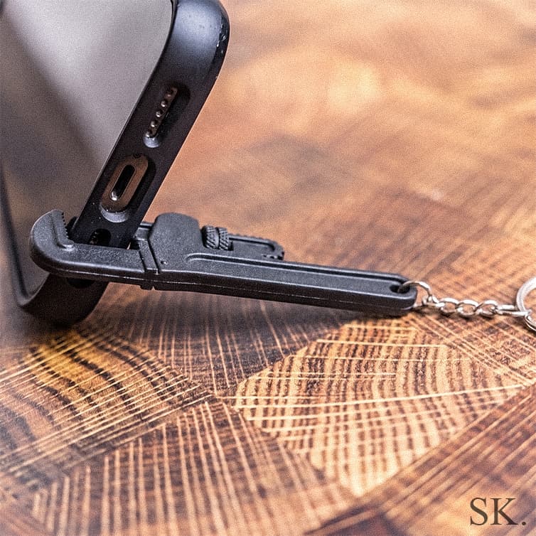 Close-up of a black miniature pipe wrench keychain supporting a smartphone as a stand, gripping its charging port on a rustic wooden table.